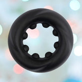The Velv'Or Rooster Cain, a black circular product crafted from soft silicone, features a ridged texture and a scalloped hole in the center, set against a softly blurred background adorned with pastel-colored lights.